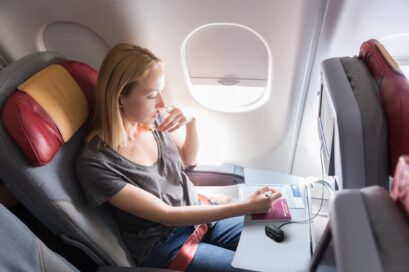 A woman sitting in the window seat of a plane is enjoying in-flight entertainment through T90S earbuds by connecting the charging case to the non-Bluetooth system