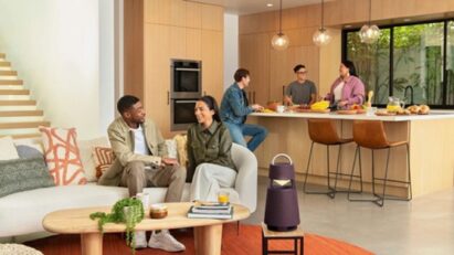 A photo of two people sitting on a couch talking and three people talking in the kitchen behind them